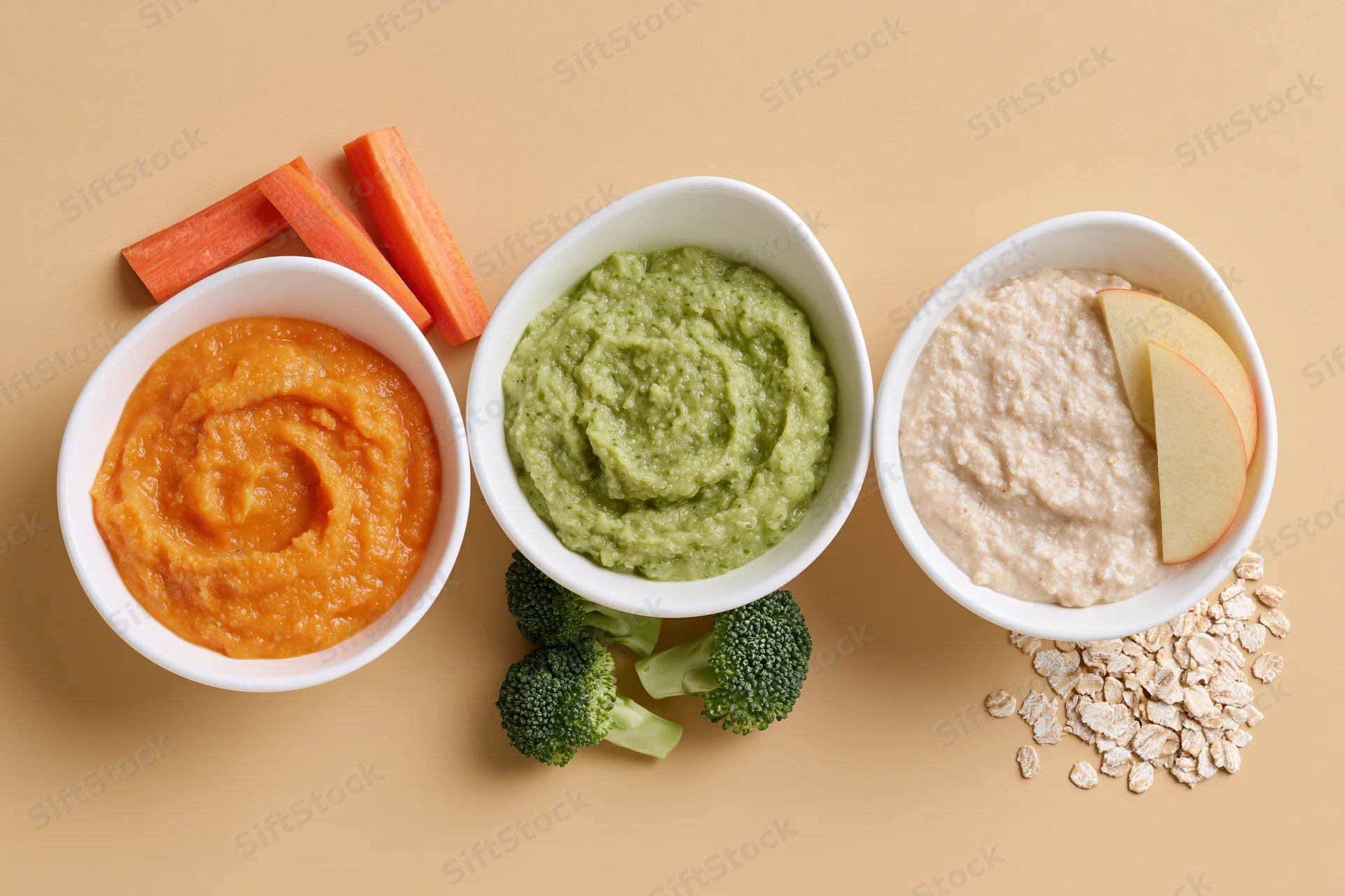 Three white ceramic bowls of baby food puree arranged in a row with fresh vegetables and fruit on beige surface