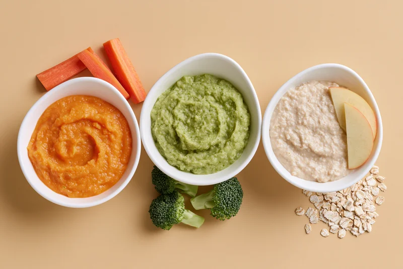 Three white ceramic bowls of baby food puree arranged in a row with fresh vegetables and fruit on beige surface
