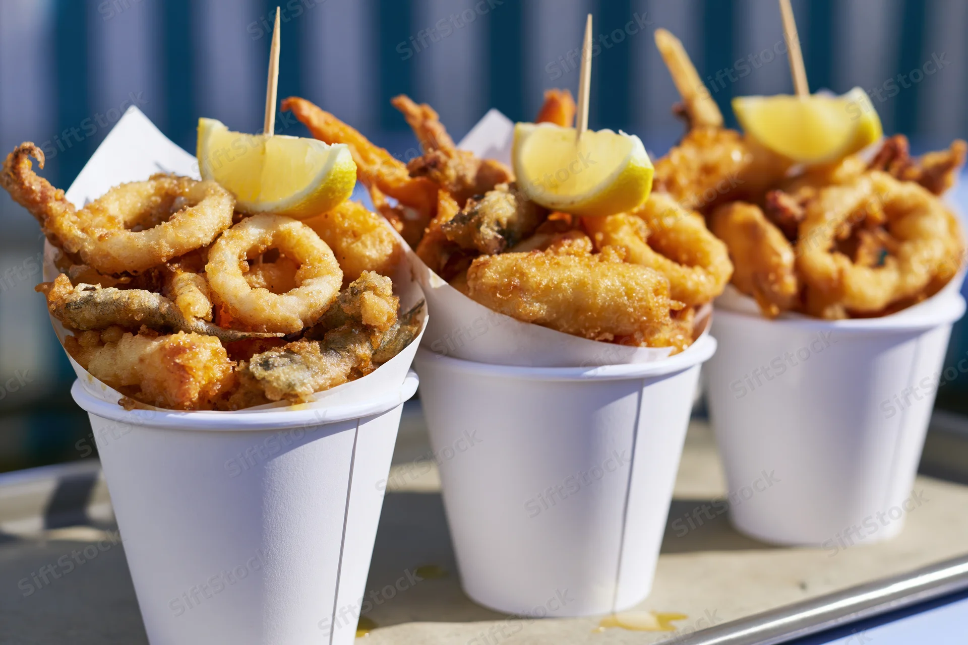 Three white paper cups filled with fried seafood and lemon wedges on a tray outdoors