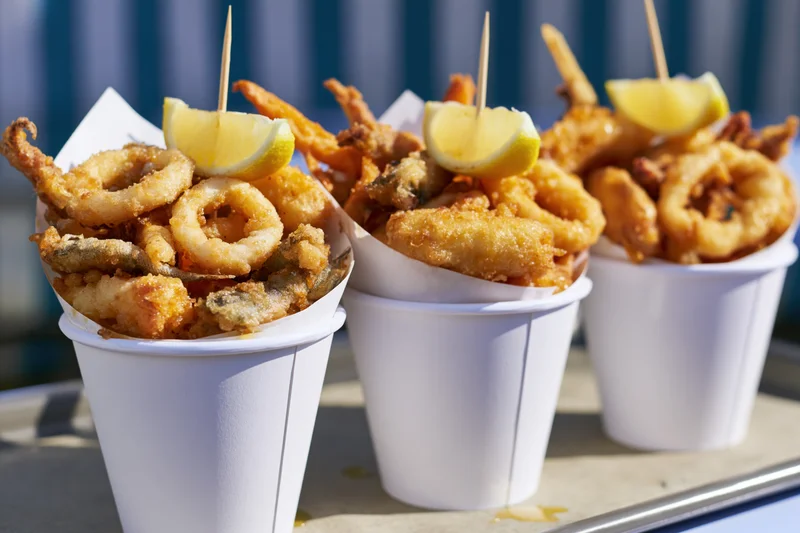 Three white paper cups filled with fried seafood and lemon wedges on a tray outdoors
