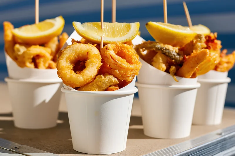 Fried seafood mix in white paper cups with lemon slices on skewers on a tray by the ocean