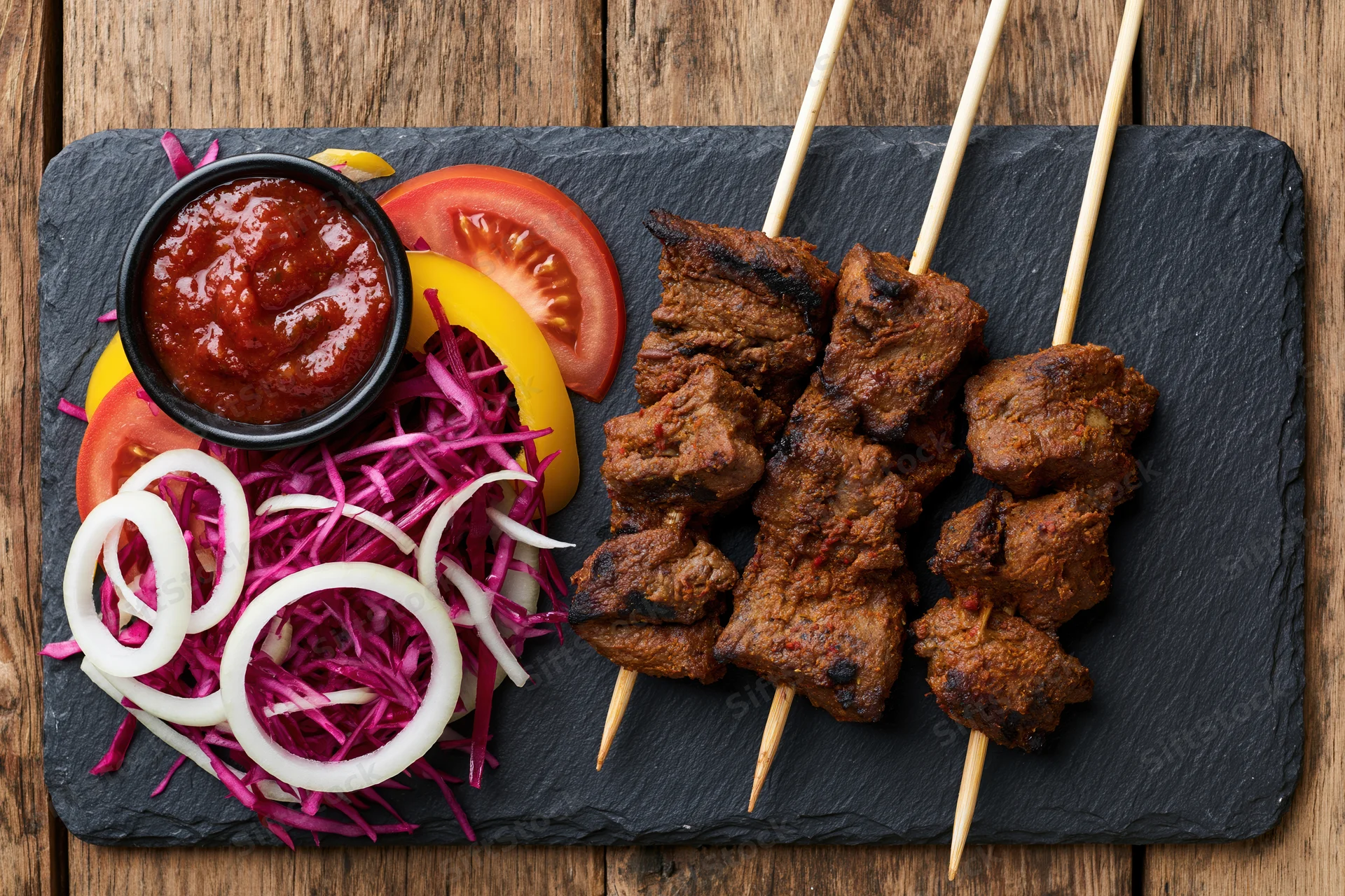Three skewers of grilled spiced meat with sauce and vegetables on a slate plate