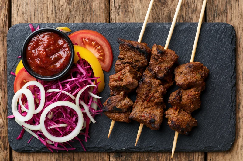 Three skewers of grilled spiced meat with sauce and vegetables on a slate plate