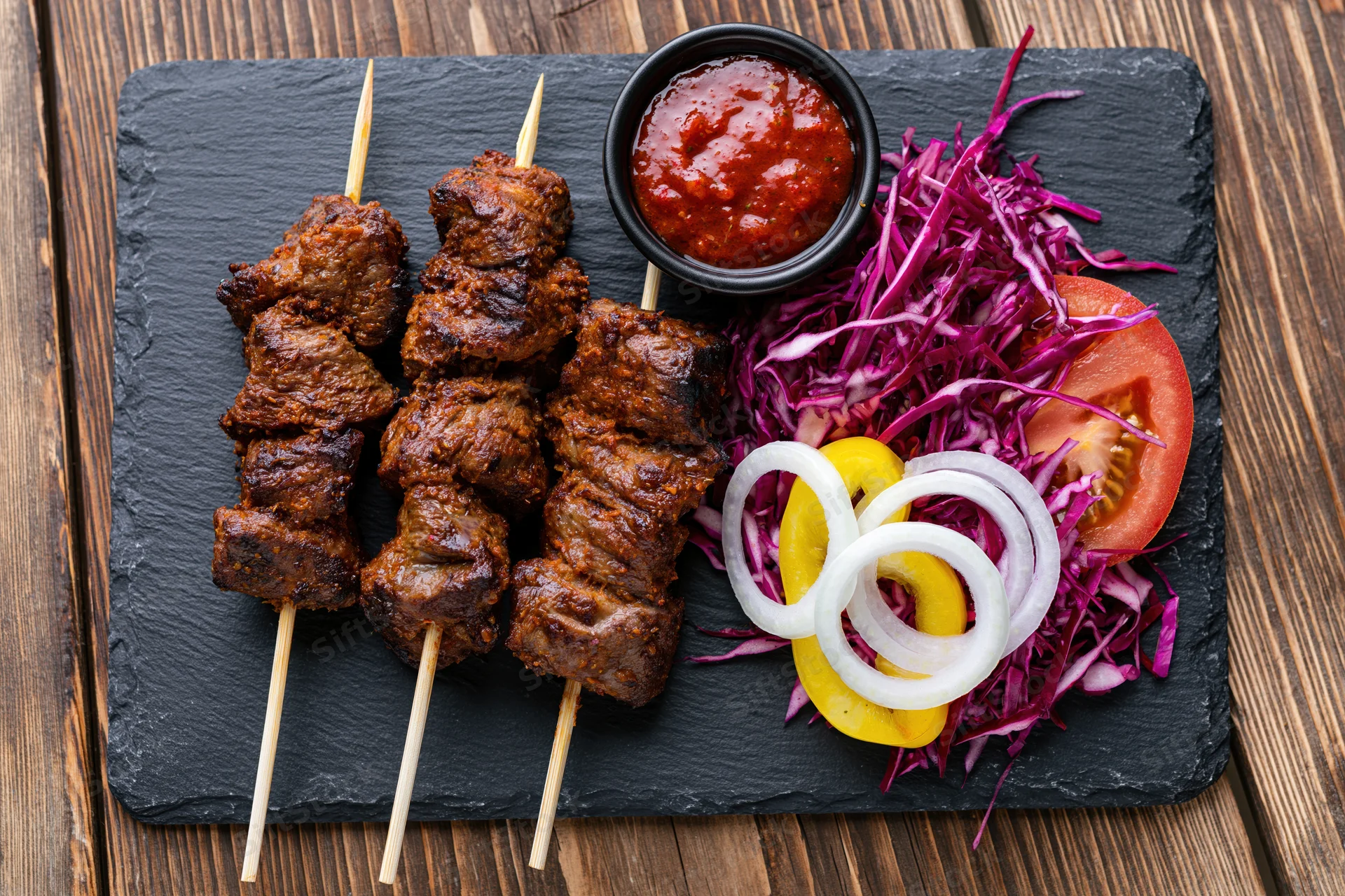 Three skewers of grilled spiced meat with sauce and vegetables on a slate plate