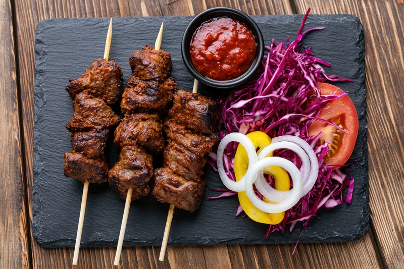 Three skewers of grilled spiced meat with sauce and vegetables on a slate plate