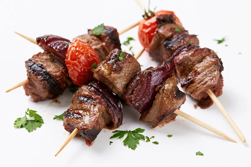 Grilled beef and tomato kebabs on wooden skewers with parsley garnish on white background