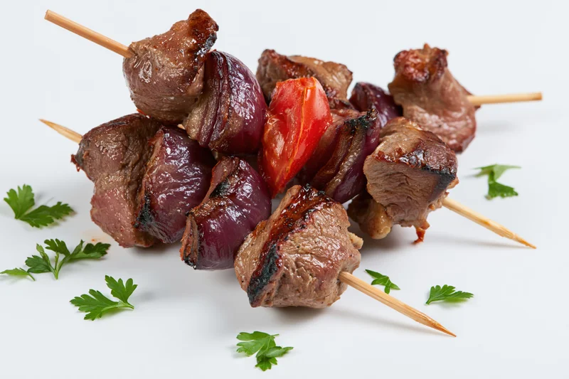 Grilled beef kebabs with red pepper and onion on wooden skewers, garnished with parsley, arranged on white background