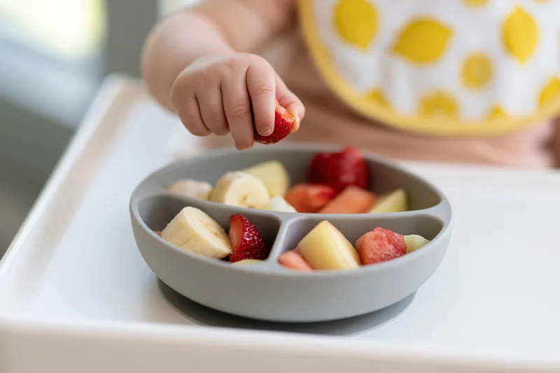 Toddler's hand picking strawberry from divided bowl of fruit on highchair tray