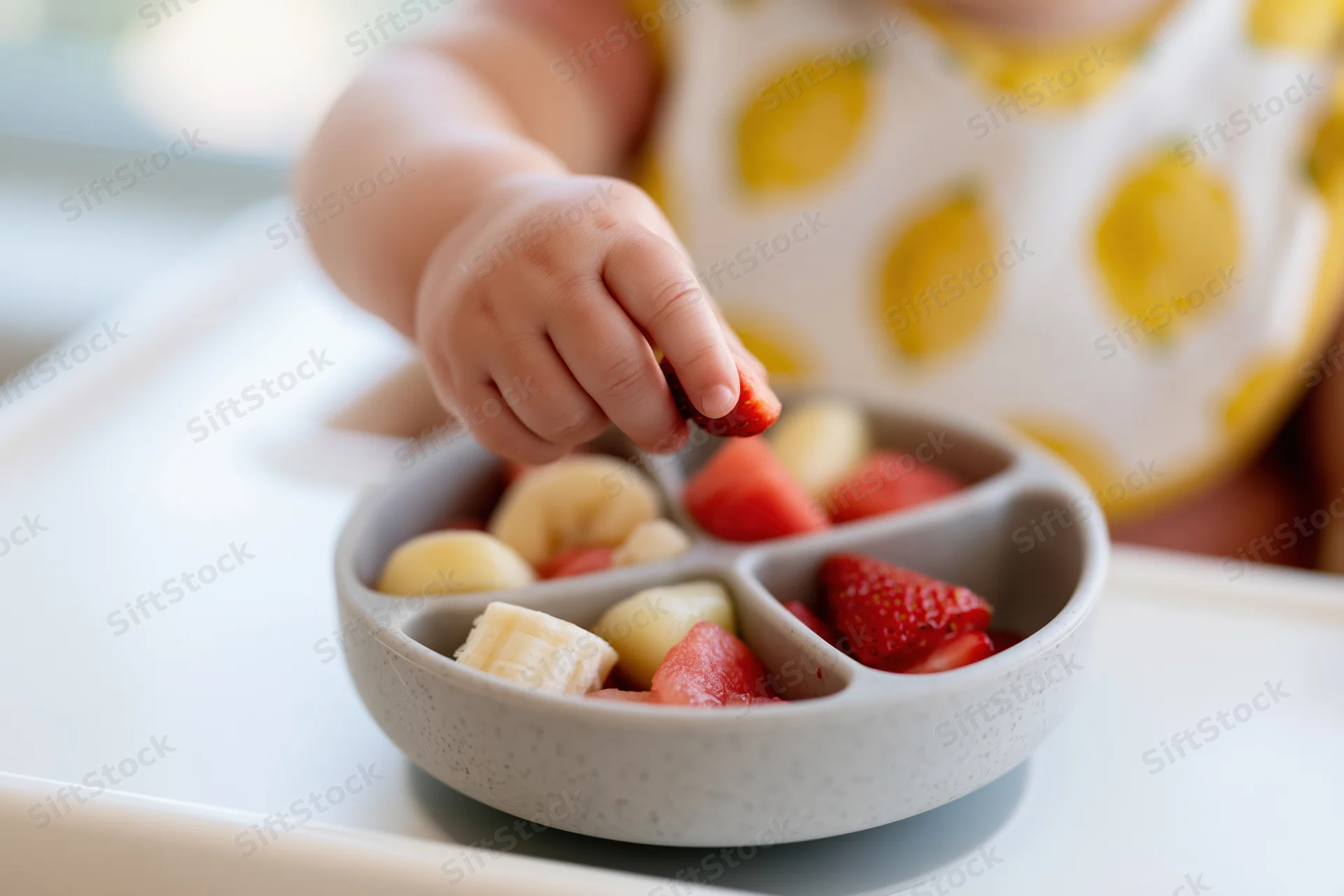 Toddler's hand picking strawberry from a divided bowl of fruit on a high chair tray