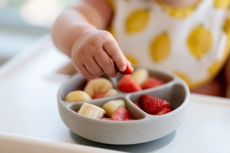Toddler's hand picking strawberry from a divided bowl of fruit on a high chair tray
