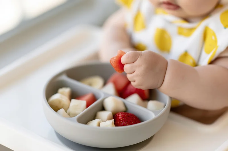 Toddler's chubby hand holding a strawberry piece over a divided bowl of fruit on a high chair tray