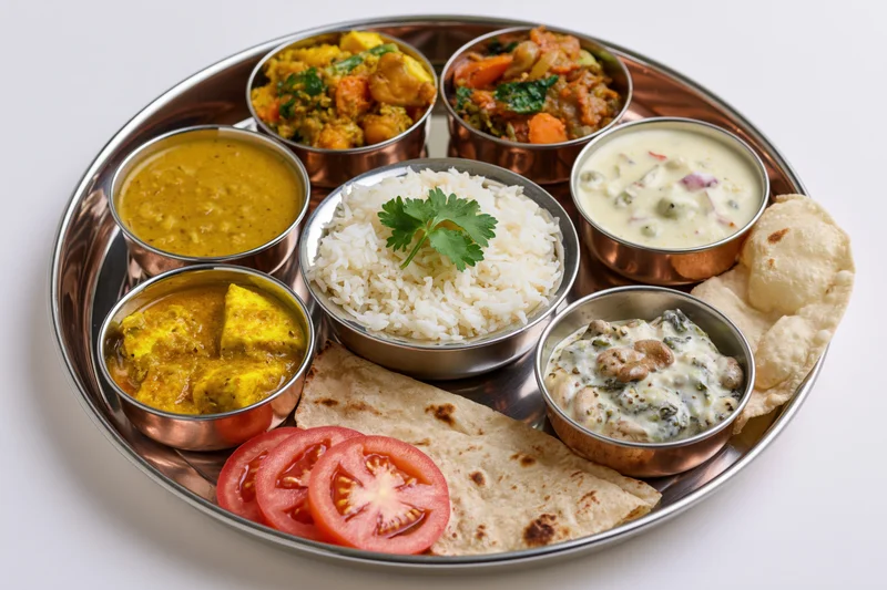 Traditional indian thali meal served on a large round metal tray with multiple small bowls of curry, rice, dal, raita, and flatbreads arranged around the center