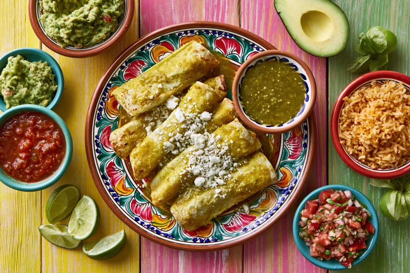 Traditional mexican enchiladas verdes served on a colorful talavera plate with rice, guacamole, salsa, and lime wedges on a painted wooden table