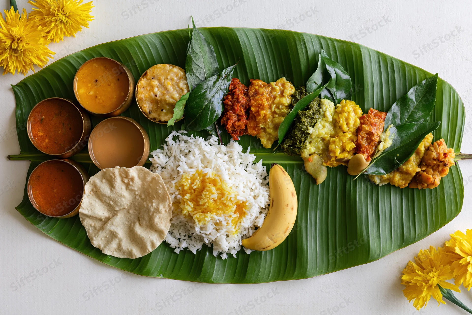 Traditional south indian onam sadya meal served on a banana leaf with rice, curries, papad, banana, and yellow flowers