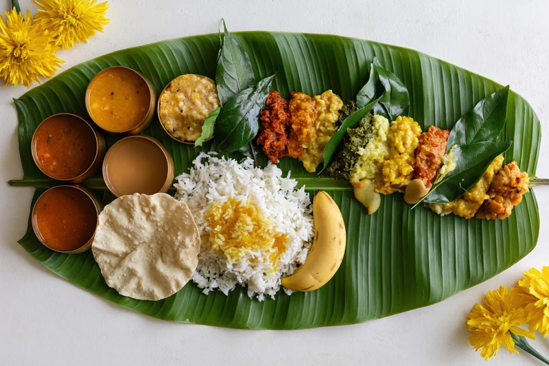 Traditional south indian onam sadya meal served on a banana leaf with rice, curries, papad, banana, and yellow flowers