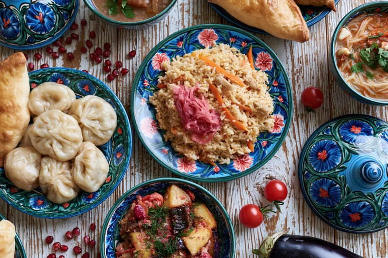 Traditional uzbeck feast spread on a rustic wooden table with patterned ceramic dishes and various dishes including pilaf, dumplings, soup, and vegetables