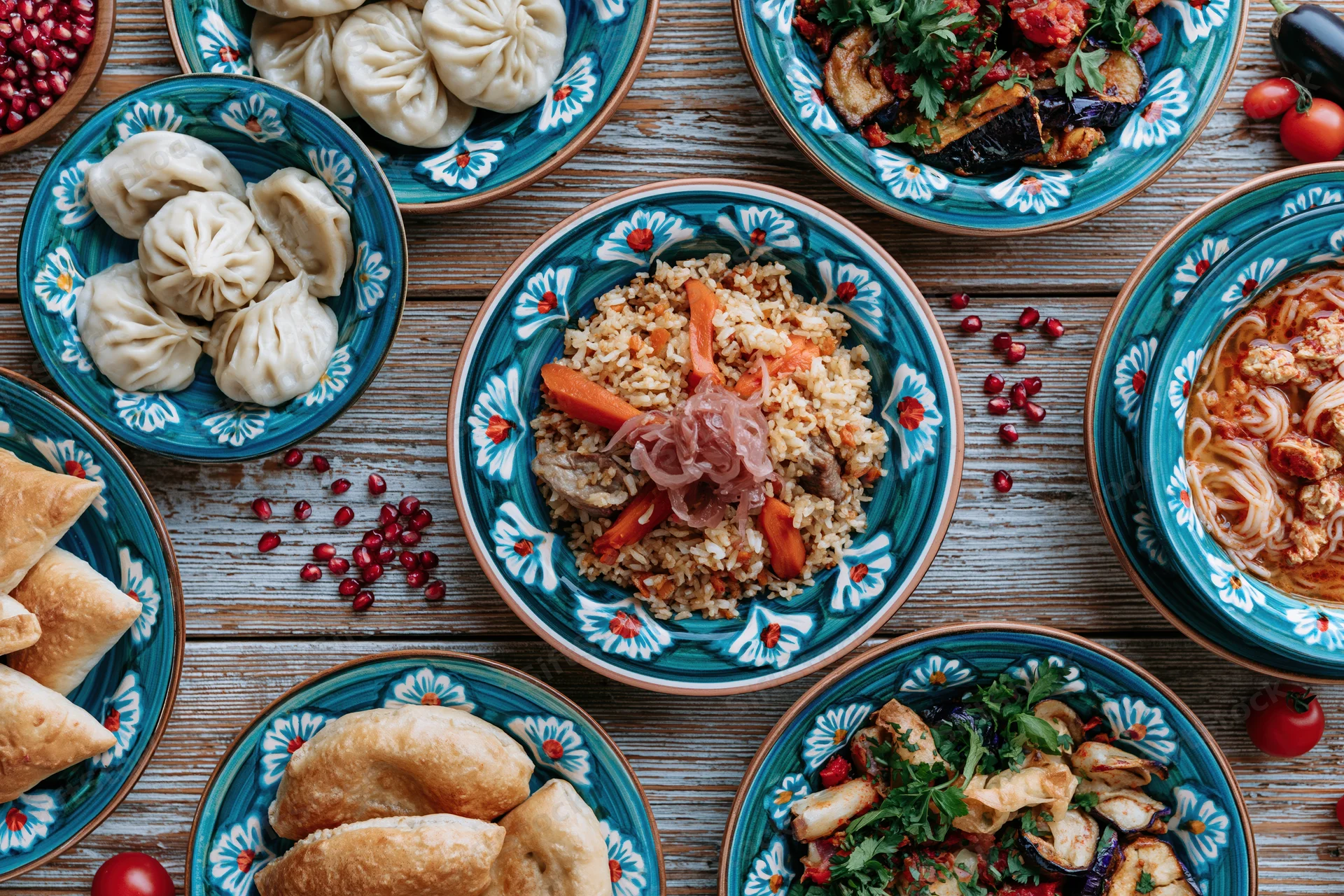 Traditional uzbe feast spread across a weathered wooden table with colorful ceramic dishes and assorted foods including pilaf, dumplings, vegetables, and pomegranate seeds