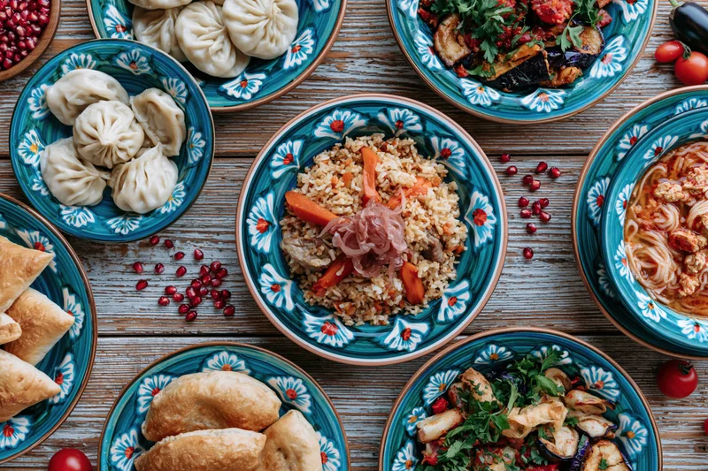 Traditional uzbe feast spread across a weathered wooden table with colorful ceramic dishes and assorted foods including pilaf, dumplings, vegetables, and pomegranate seeds