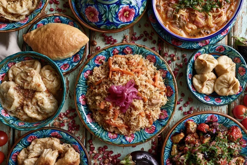 Traditional uzbe feast spread across a weathered wooden table with colorful ceramic dishes filled with pilaf, dumplings, soup, bread and vegetables