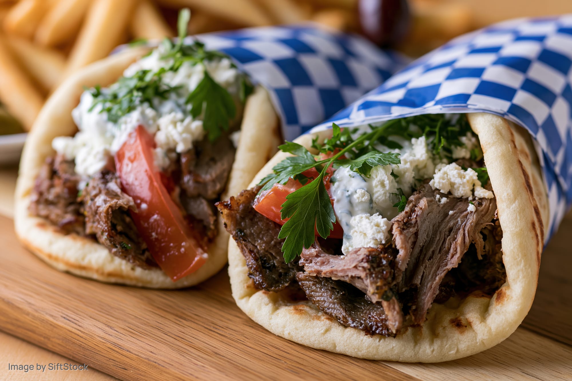 Two Greek gyros wrapped in checkered paper on a wooden cutting board with fries in the background