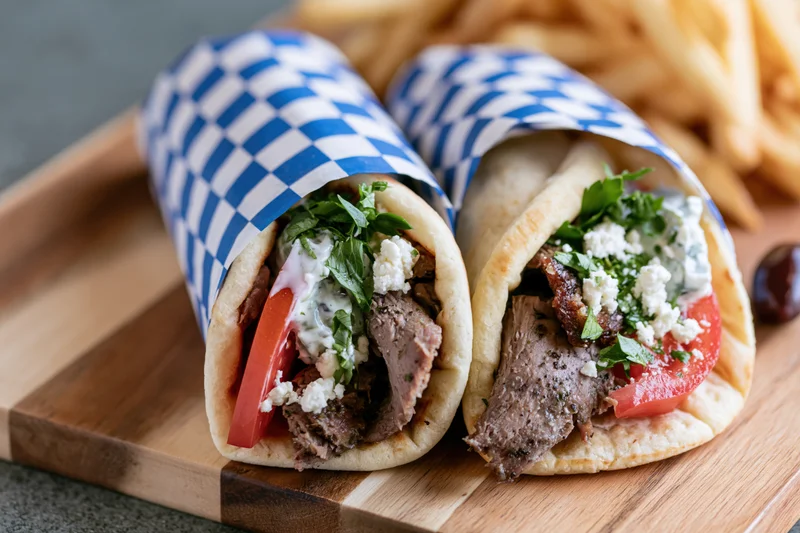 Two Greek gyros wrapped in checkered paper on a wooden cutting board with fries in the background