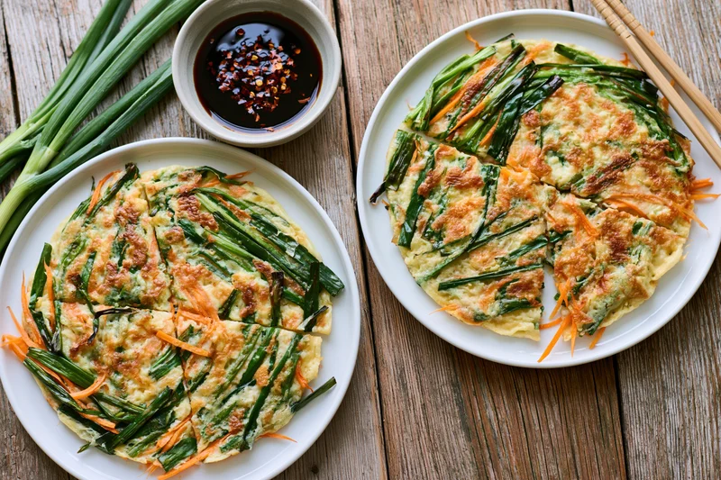 Two korean scallion pancakes served on white plates with dipping sauce and chopsticks on a wooden table