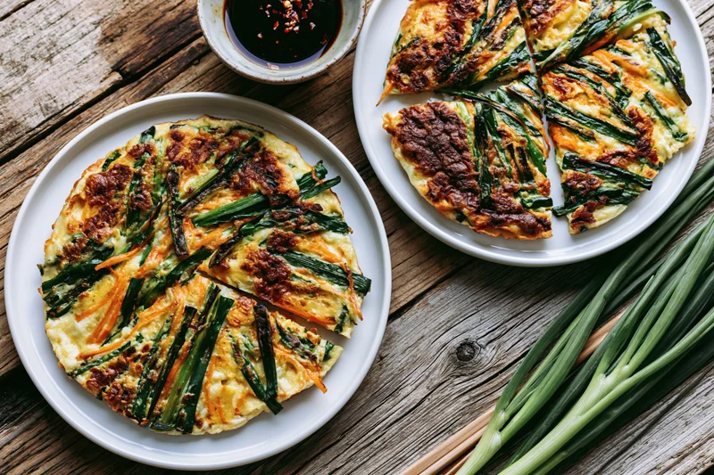 Two korean scallion pancakes served on white plates with dipping sauce and fresh green onions on a rustic wooden table