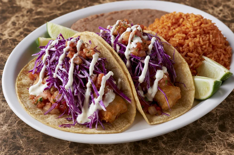 Two battered fish tacos in soft corn tortillas with purple cabbage and white sauce on a white plate with red rice, refried beans, and lime wedges