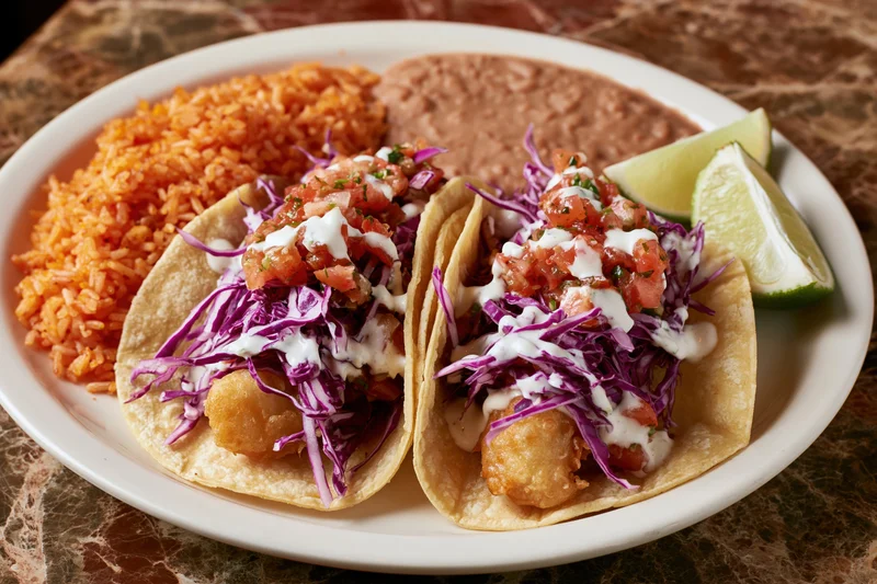 Two battered fish tacos in soft corn tortillas topped with purple cabbage, salsa, and sour cream on a white plate with red rice, refried beans, and lime wedges