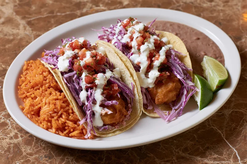Two battered fish tacos in soft corn tortillas with red cabbage, sour cream, pico de gallo, and lime wedges on a white plate with red rice and refried beans on a marble table