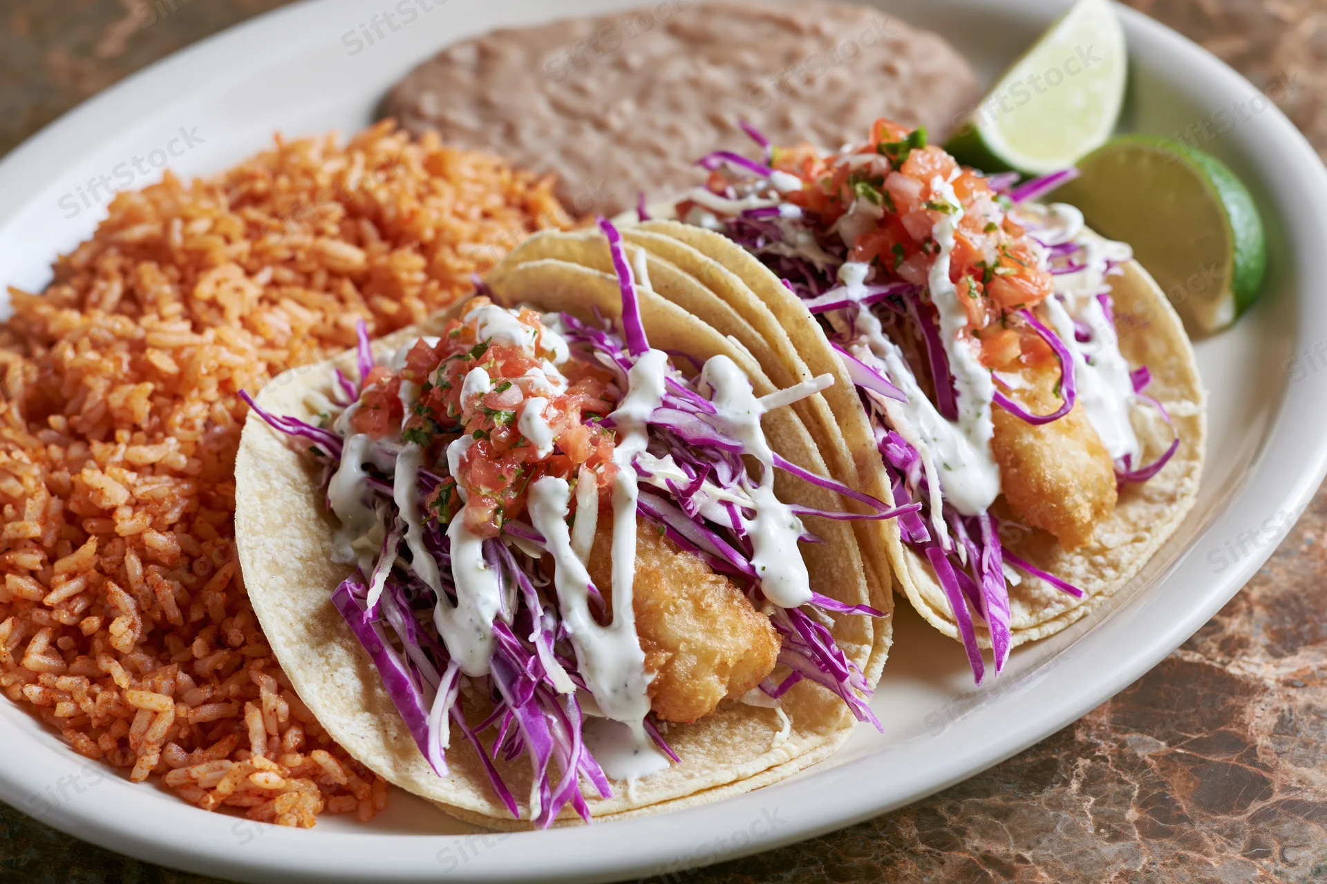 Two battered fish tacos in soft corn tortillas with purple cabbage and white sauce served with red rice and refried beans on a white plate