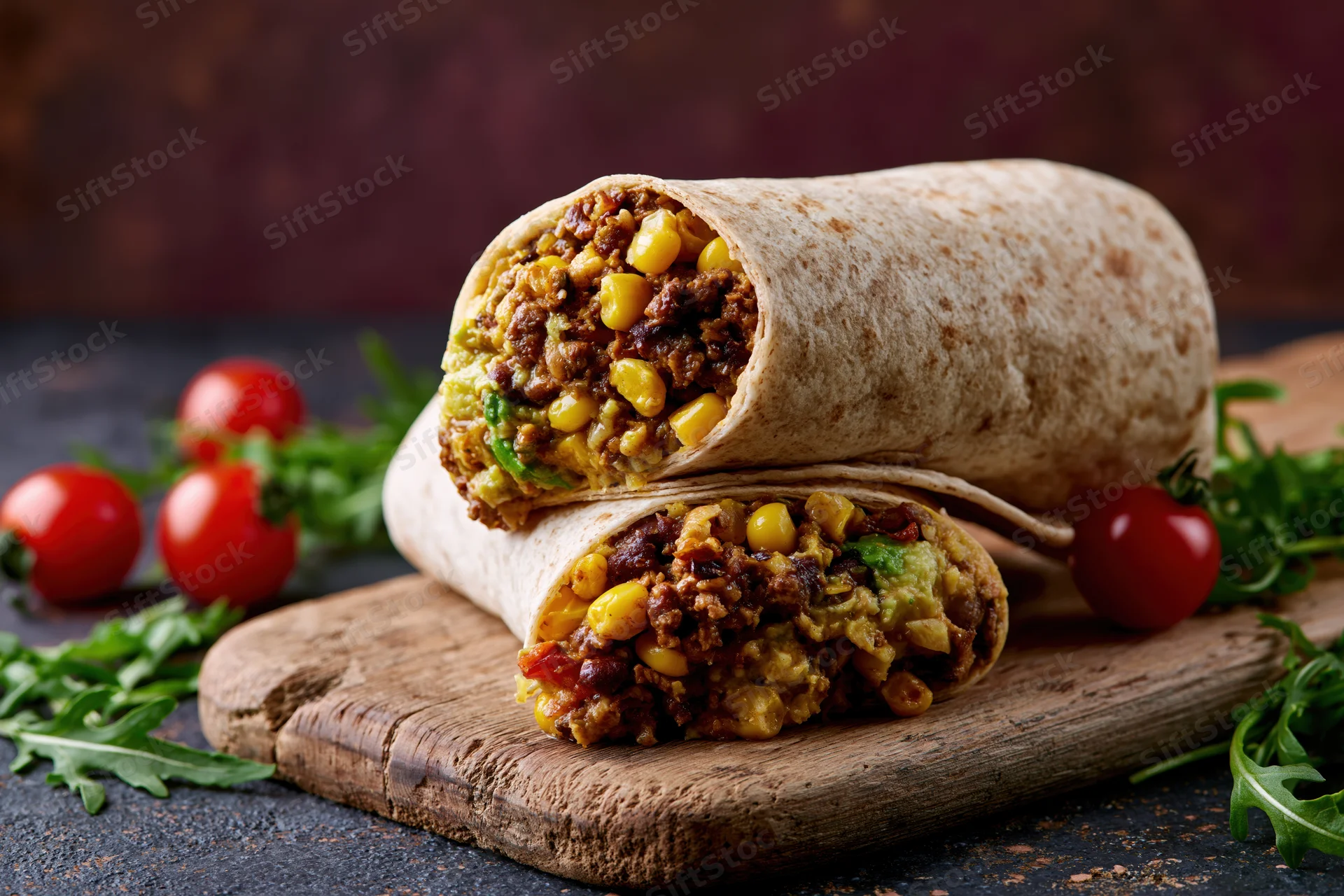 Two beef and vegetable burritos cut in half and stacked on a wooden cutting board with cherry tomatoes and arugula