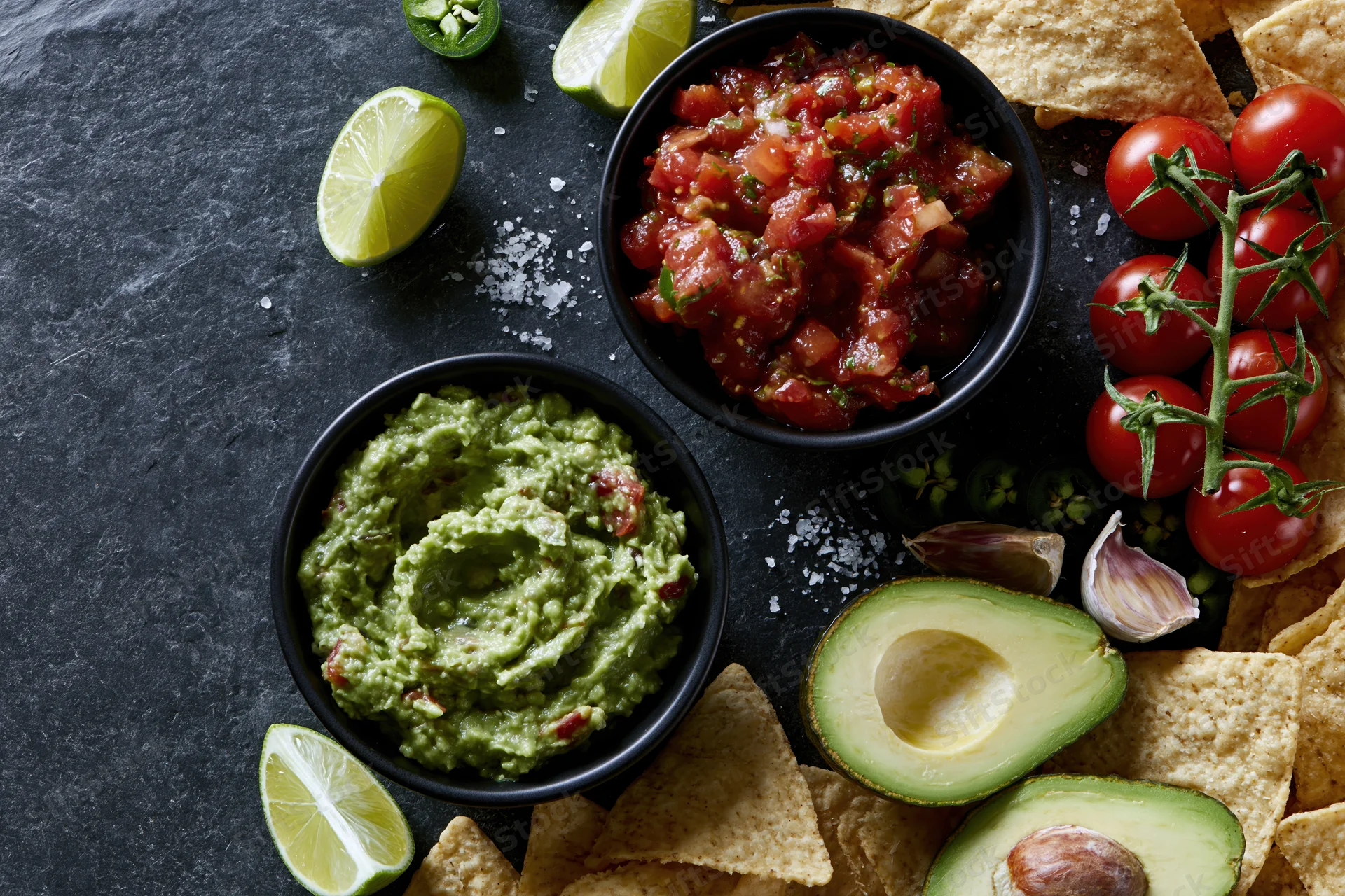 Two black bowls of guacamole and fresh tomato salsa surrounded by avocado halves, lime wedges, tortilla chips, cherry tomatoes on vine, garlic, jalapeno, and salt on dark slate surface