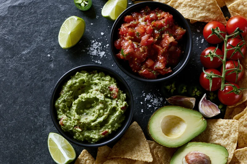 Two black bowls of guacamole and fresh tomato salsa surrounded by avocado halves, lime wedges, tortilla chips, cherry tomatoes on vine, garlic, jalapeno, and salt on dark slate surface