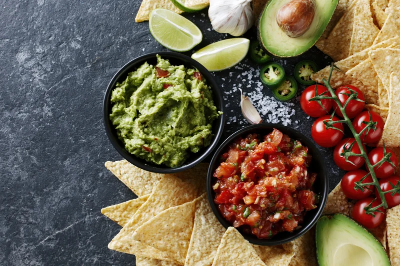 Guacamole and salsa in black bowls surrounded by tortilla chips and fresh ingredients on dark stone surface
