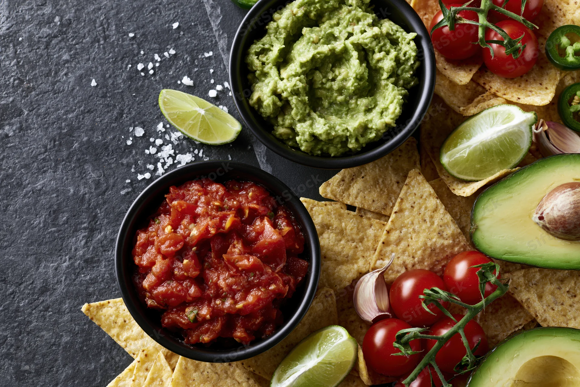 Guacamole and salsa in black bowls with tortilla chips, avocado, lime, tomatoes, and garlic on a dark stone surface