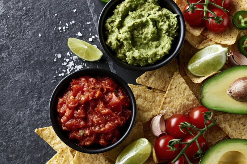 Guacamole and salsa in black bowls with tortilla chips, avocado, lime, tomatoes, and garlic on a dark stone surface