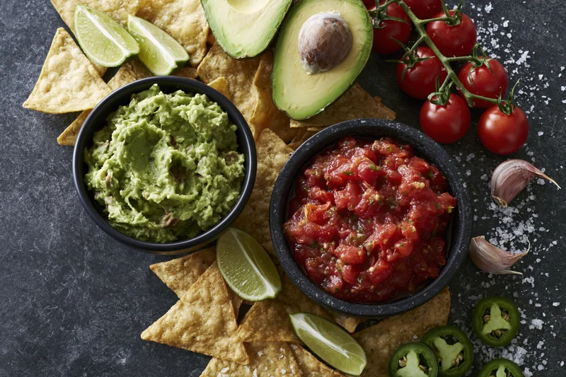 Two black bowls of guacamole and fresh tomato salsa surrounded by tortilla chips, avocado, lime, garlic, jalapeno, and salt on a dark stone surface