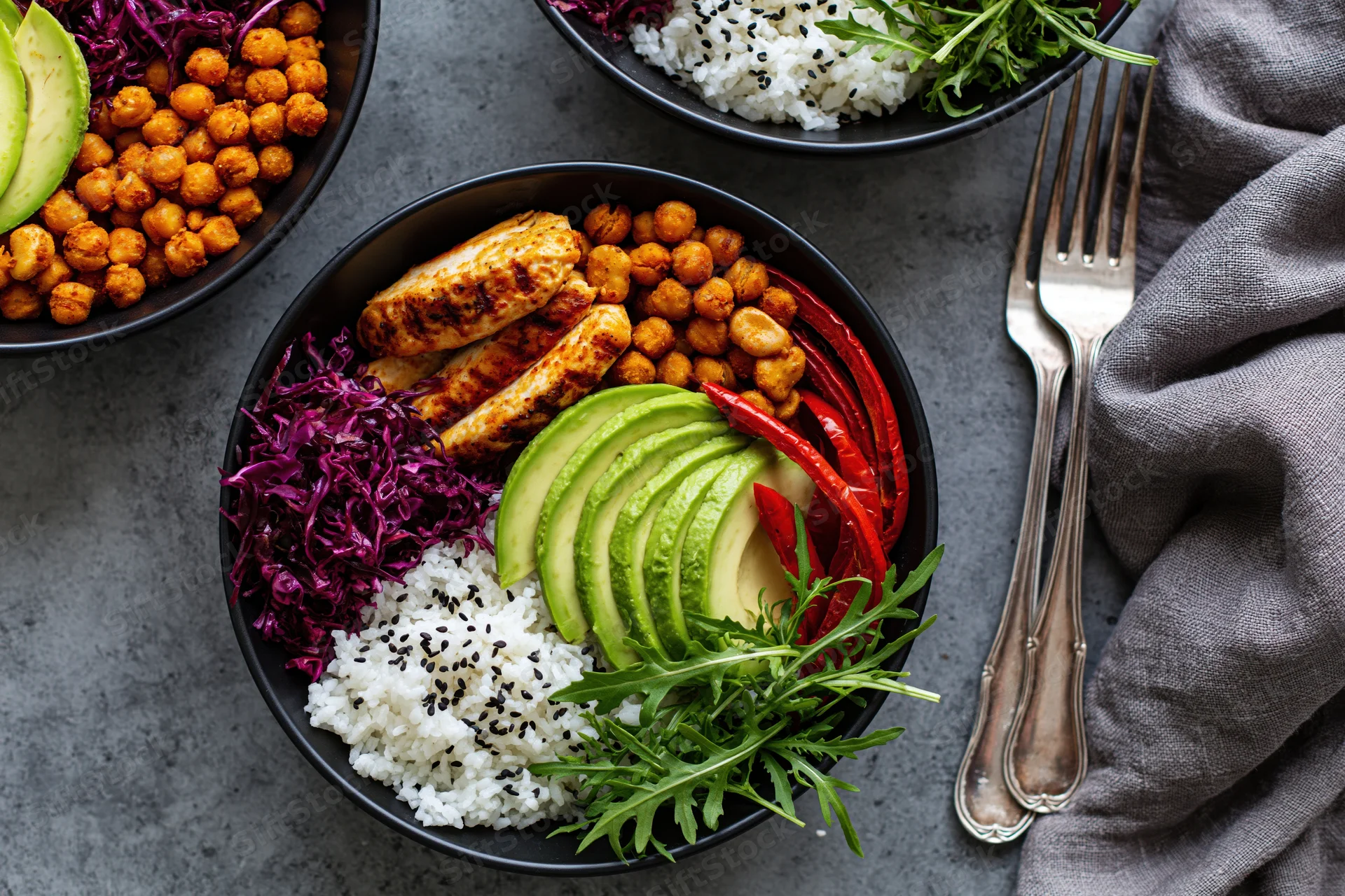 Two black bowls with grilled chicken, avocado, rice, chickpeas, red cabbage, arugula and peppers on a gray table with forks and a napkin