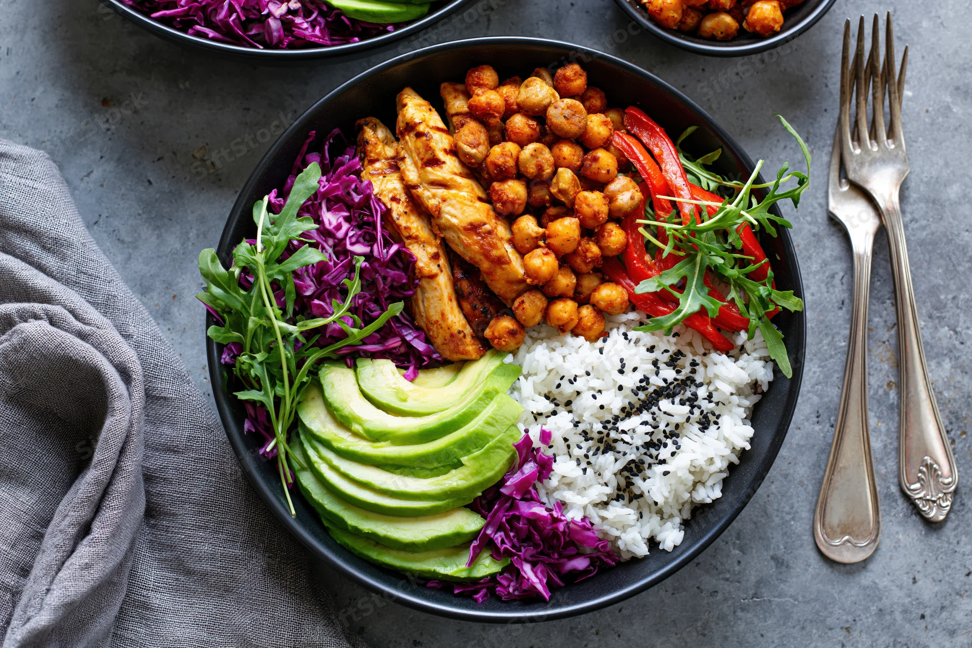 Black bowl filled with grilled chicken, rice, avocado, chickpeas, purple cabbage and arugula on gray surface with forks and cloth napkin