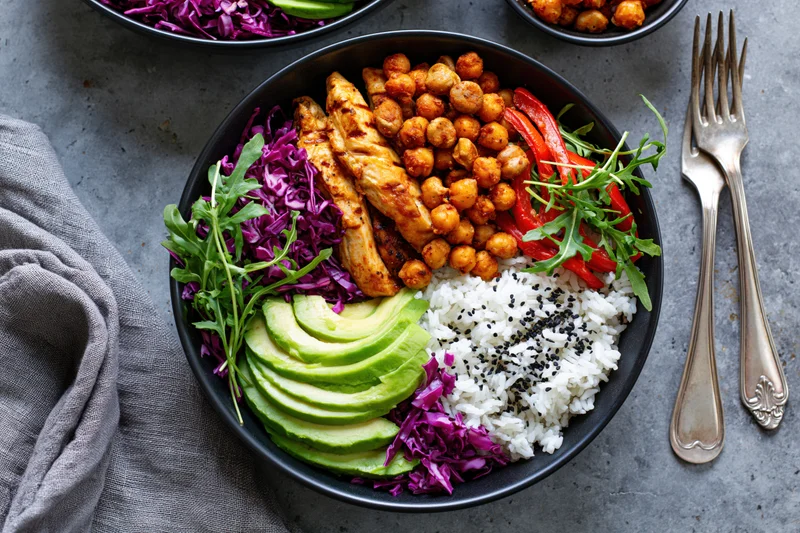 Black bowl filled with grilled chicken, rice, avocado, chickpeas, purple cabbage and arugula on gray surface with forks and cloth napkin