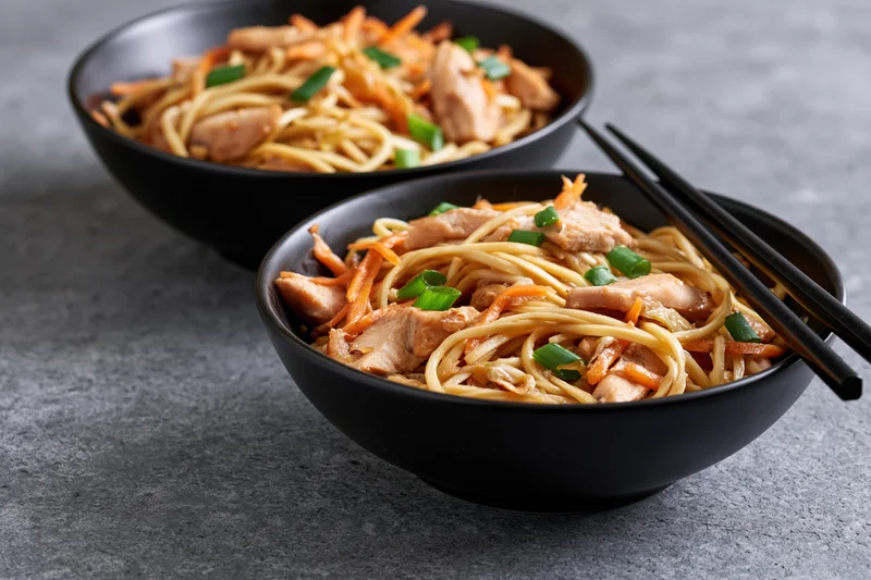 Two black bowls of stir-fried egg noodles with chicken and vegetables on a gray stone surface