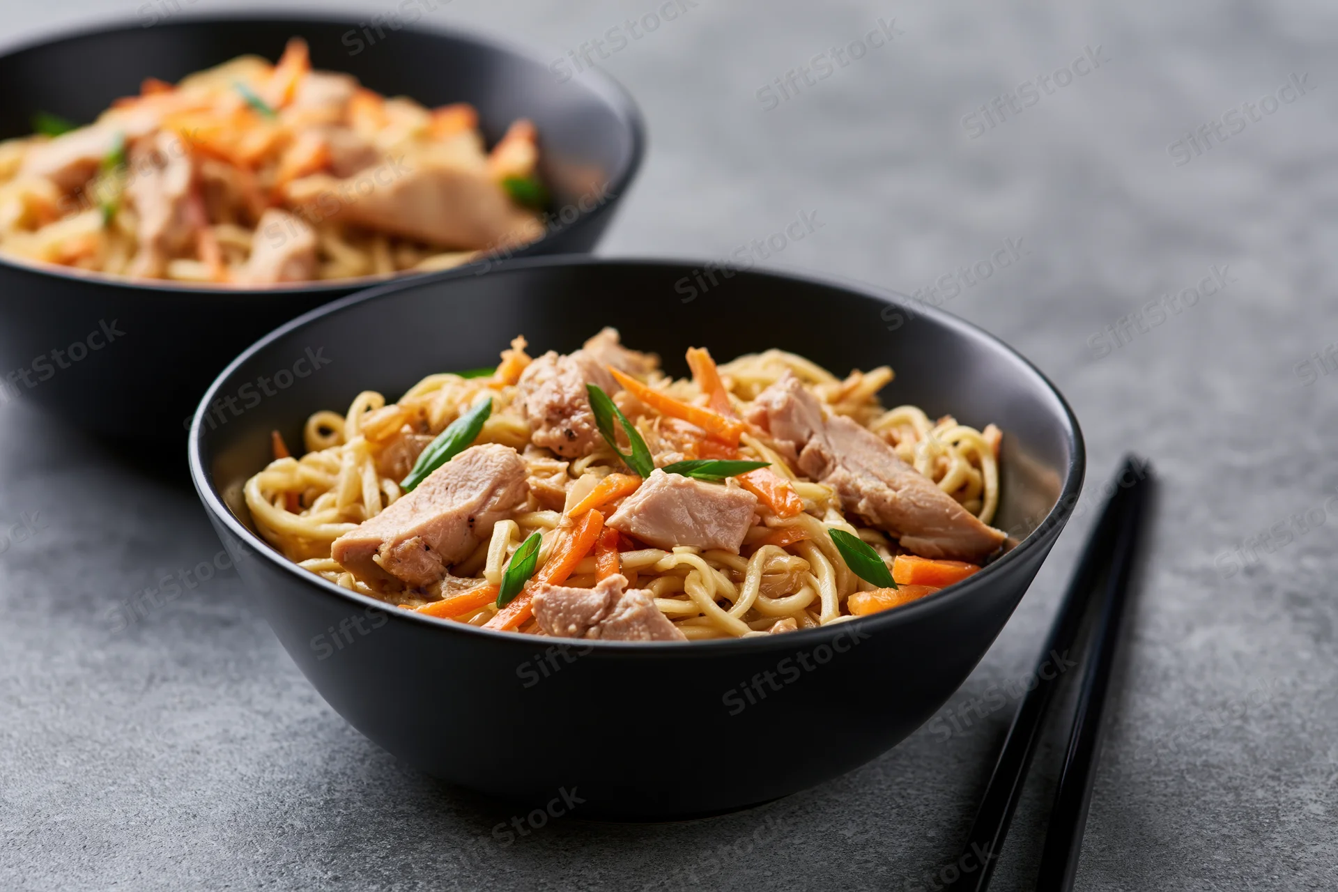 Two black bowls of stir-fried egg noodles with chicken and vegetables on a gray surface, chopsticks beside
