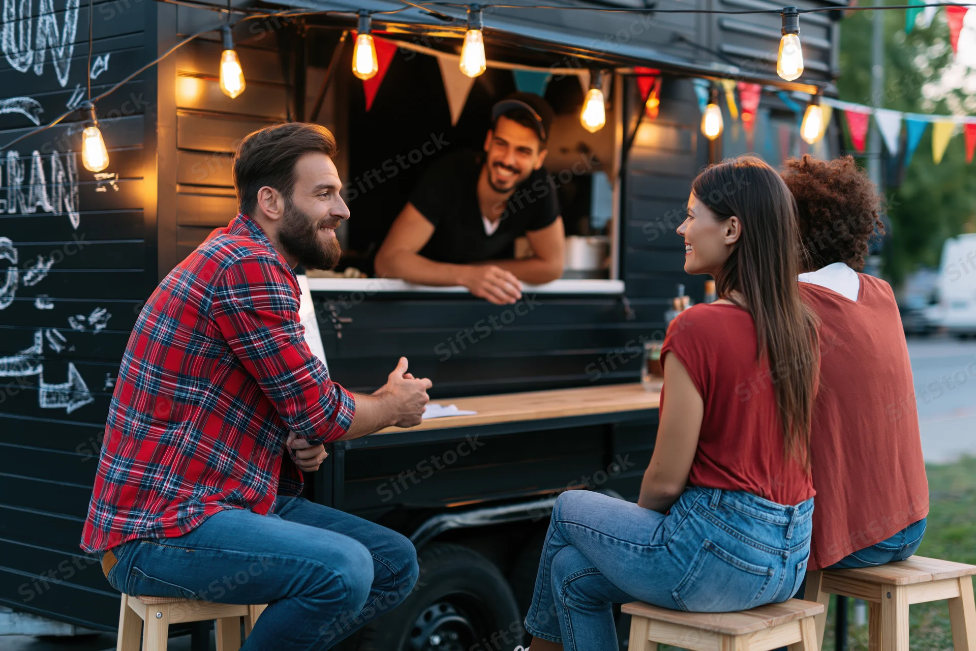 Two customers chatting and ordering at a black food truck counter outdoors in the evening