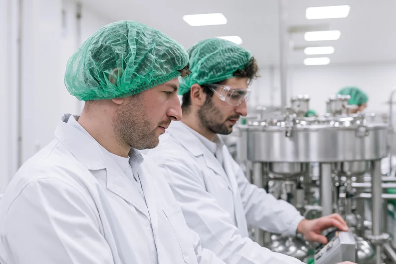 Two men in white lab coats and green hairnets operating machinery in a cleanroom facility