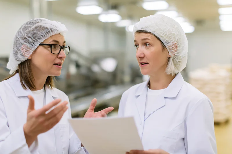 Two women in white lab coats and hairnets discussing a document in a food factory setting