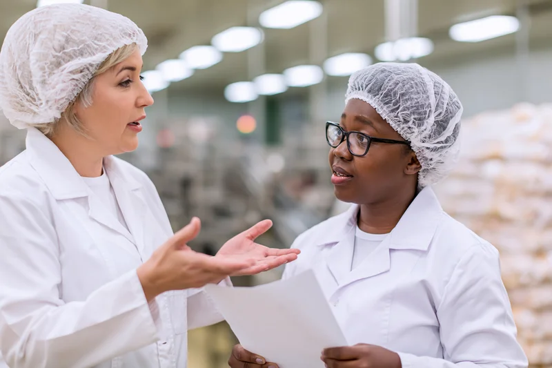 Two women in lab coats and hairnets discussing a document in a food factory