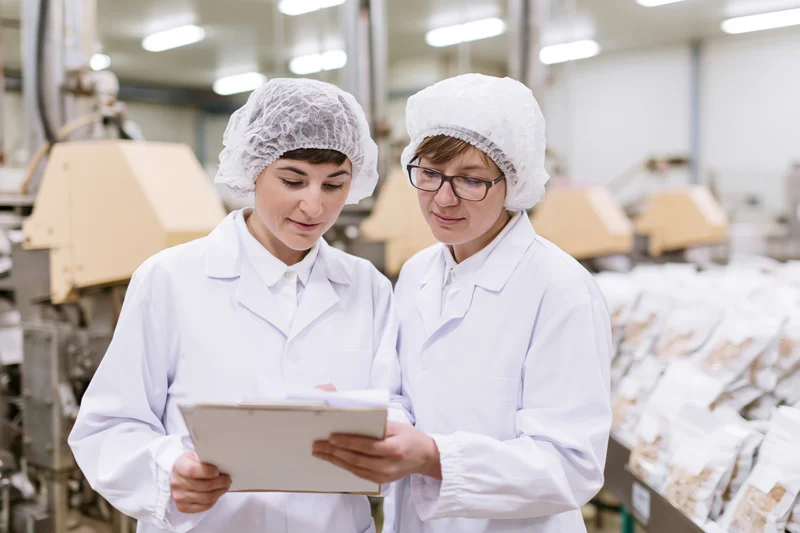 Two women in lab coats and hairnets reviewing a clipboard in a food factory production area