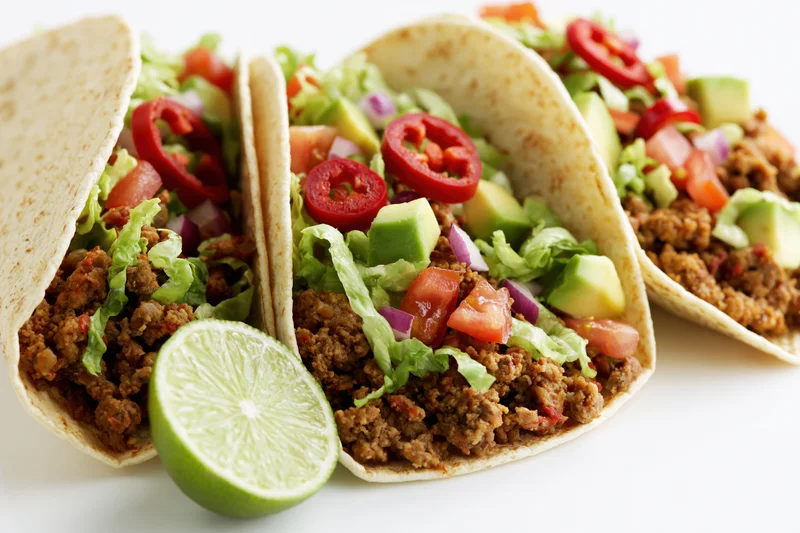 Three tacos filled with seasoned ground beef and topped with lettuce, tomato, avocado, jalapeno, and lime on a white surface