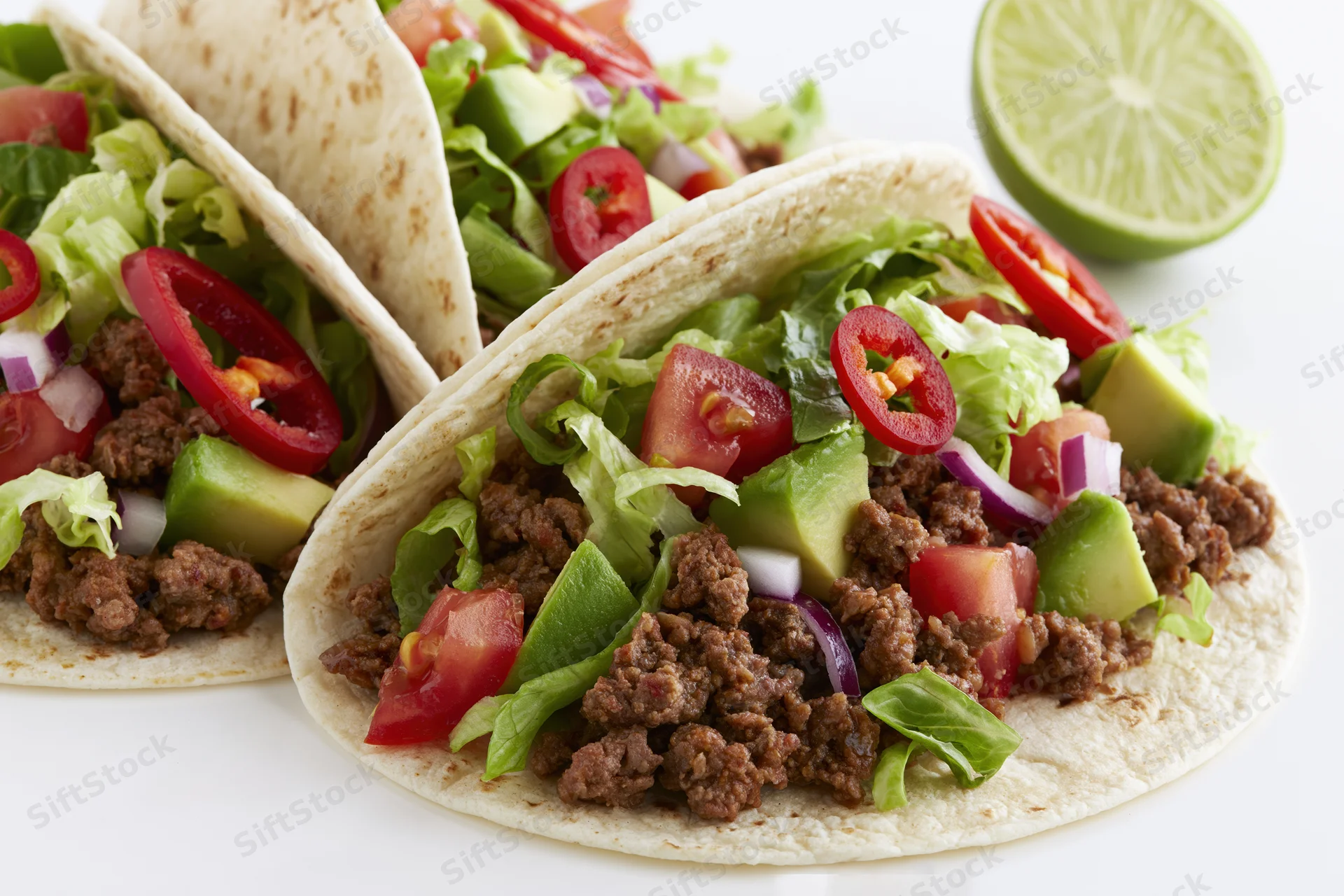 Two flour tortilla tacos filled with seasoned ground beef and topped with lettuce, tomato, avocado, red onion, and sliced chili, with a lime wedge on the side, on a white surface